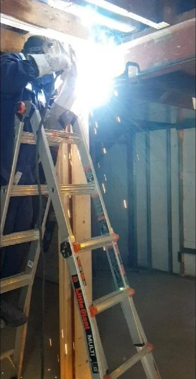 A welder in a helmet welding overhead from a ladder, sparks lighting the room